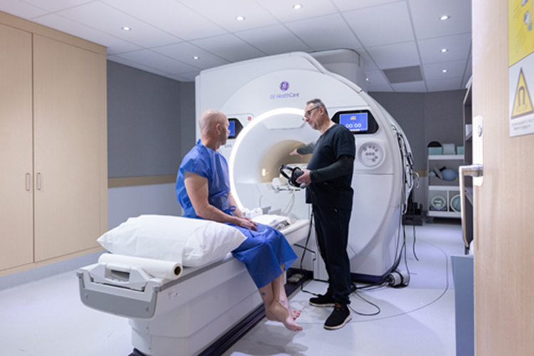Man sitting on bed of MRI machine with radiographer explaining scan to him.