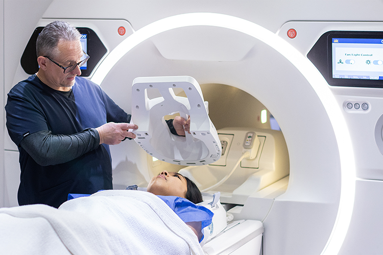Male radiographer placing imaging equipment over female patient's head before MRI to help increase image quality.